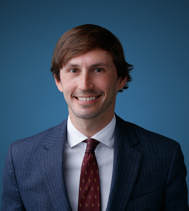 Professional headshot of a smiling man in a navy pinstripe suit, white shirt, and burgundy tie against a blue background.